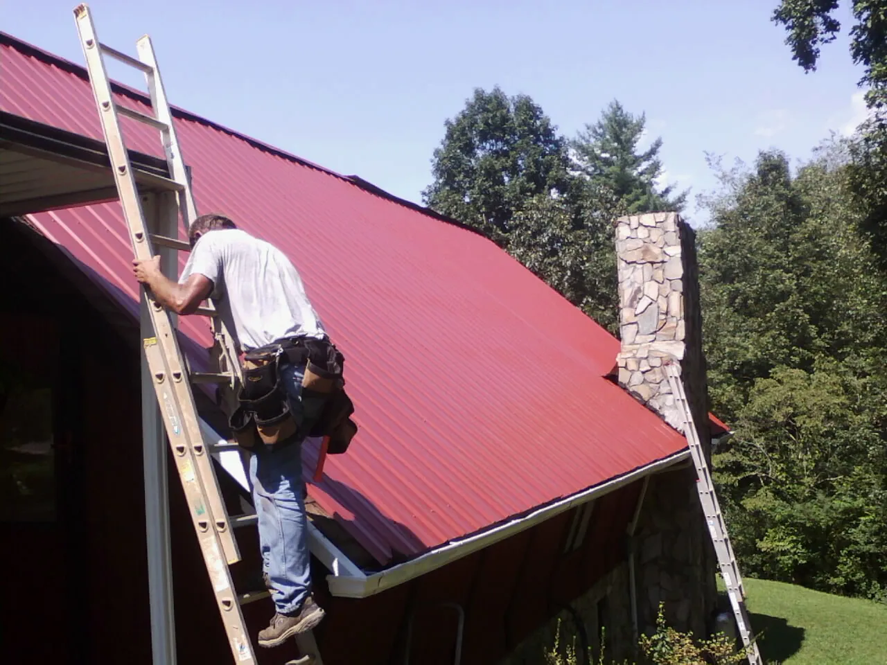 Roofer on a ladder beside a red metal roof and stone chimney — Soffit & Fascia Repair by Roofing Brothers in South Greenfield